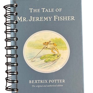 May include: A spiral-bound book with a blue-gray cover featuring the title "The Tale of Mr. Jeremy Fisher" in white text. A circular illustration depicts a frog fishing. The author's name, Beatrix Potter, is also printed on the cover.