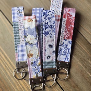 May include: A collection of fabric key fobs with various floral and patterned designs. The key fobs feature a mix of colors, including blue, pink, and white, and are attached to metal key rings. The key fobs are arranged on a wooden surface.