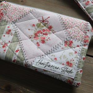 May include: Quilted floral clutch with a matching wrist strap. The patchwork design features pink and green floral patterns on a light background. The clutch has a zipper closure and a small tag that reads "Heartwoven Studio."