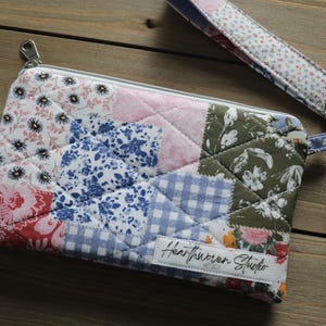 May include: A quilted patchwork clutch with a wrist strap, featuring a variety of floral and geometric patterns in shades of pink, blue, green, and white. The clutch has a silver zipper and a label that reads "Heartwoven Studio."