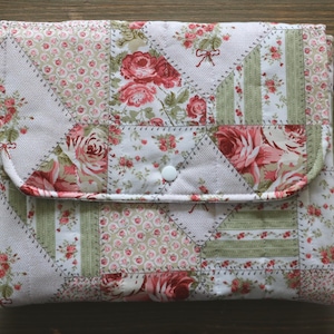 May include: A quilted fabric case with a flap closure. The case features a patchwork design with floral patterns in shades of pink, red, and green on a white background. A white button secures the flap. The case is likely for a tablet or e-reader.