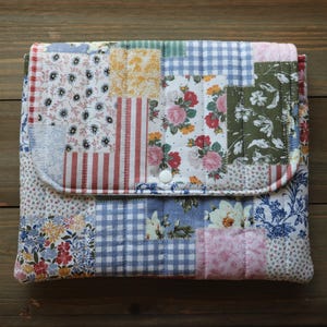 May include: A patchwork fabric case with a flap closure. The case features a variety of floral, striped, and gingham patterns in shades of red, blue, pink, green, and yellow. The case is secured with two white buttons.