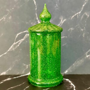 May include: A green glitter-covered jar with a lid and a pointed finial. The jar has a cylindrical body and a decorative base. The lid is flat with a glittery surface. The jar is displayed against a dark background.