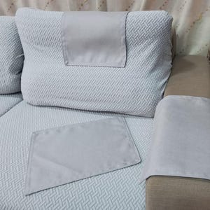 May include: Light gray sofa cushions and armrest covers. The cushions have a textured, woven pattern. The armrest cover is a solid light gray color. The sofa is a neutral color, with a light gray patterned fabric.