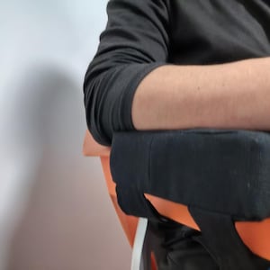 Puede incluir: Cojín negro para reposabrazos sujeto a una silla naranja. El cojín parece estar hecho de una tela suave y texturizada. La silla es parcialmente visible, con el reposabrazos posicionado para dar soporte.