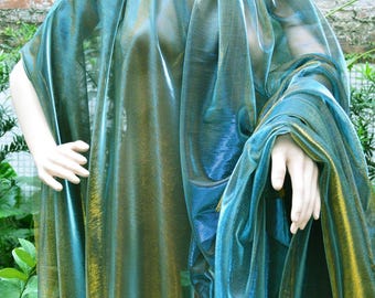 May include: A flowing, iridescent teal and gold shawl draped over a mannequin. The fabric has a shimmering, semi-transparent quality, with a gradient of colors. The garment appears to be lightweight and suitable for layering or as an accessory.