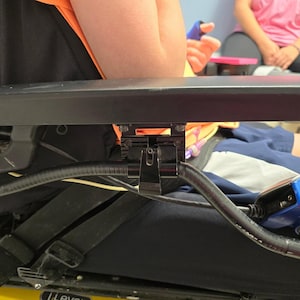 May include: A black armrest with a metal bracket attached to a wheelchair. The armrest is adjustable and can be moved up and down.