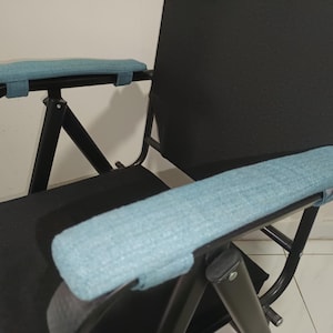 Puede incluir: Una silla plegable negra con reposabrazos acolchados azules. La silla tiene un marco de metal negro y un asiento de tela negro.