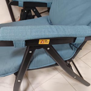 Könnte beinhalten: Ein blauer Klappstuhl mit einem schwarzen Metallrahmen und gepolsterten Armlehnen. Der Stuhl hat ein Warnschild mit der Aufschrift "Warnung: Nicht auf dem Stuhl stehen."