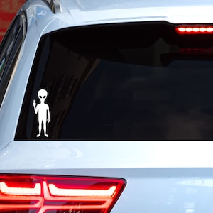 May include: A white alien sticker on the back window of a white car. The alien is giving the middle finger.