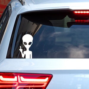 Puede incluir: Una pegatina de un extraterrestre blanco con un dedo medio en un coche plateado. La pegatina está en la ventana trasera del coche.
