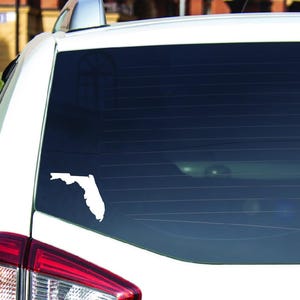 Puede incluir: Pegatina de vinilo blanco con el contorno del estado de Florida en la ventana trasera de un coche blanco.