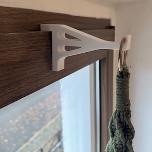 Könnte beinhalten: Weißer, 3D-gedruckter Pflanzenaufhänger an einer Holzoberfläche befestigt. Der Halter trägt einen grünen Makramee-Pflanzenhalter mit einem goldfarbenen Ring. Der Halter hat eine dreieckige Form mit drei Aussparungen.