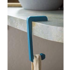 Könnte beinhalten: Ein türkisblauer Türhaken mit einem beigen Handtuch. Der Haken besteht aus einem soliden Material und hat ein schlichtes, modernes Design. Das Handtuch ist hellbeige und aus einem weichen Stoff gefertigt.