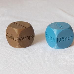 Könnte beinhalten: Zwei Würfel mit verschiedenen Speisenoptionen, die auf jeder Seite aufgedruckt sind. Ein Würfel ist braun mit den Worten "Auflauf", "Wraps", "Grillen", "Pizza", "Salat" und "Pasta". Der andere Würfel ist blau mit den Worten "Burger", "Döner", "Pizza", "Pasta", "Salat" und "Wraps".