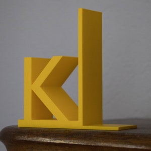 Könnte beinhalten: Ein gelber, plastischer Buchstütze in Form des Buchstabens "K" mit einem vertikalen Stück, das an der rechten Seite befestigt ist.