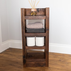 May include: A dark brown wooden shelf unit with four shelves. The top shelf has a glass bottle with wooden sticks in it. The second shelf has two towels, one brown and one gray. The third shelf has two rolls of toilet paper. The bottom shelf is empty.