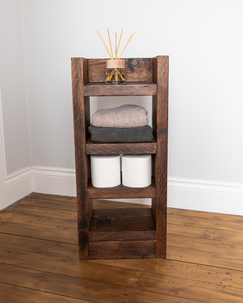 May include: A dark brown wooden shelf unit with four shelves. The top shelf has a glass bottle with wooden sticks in it. The second shelf has two towels, one brown and one gray. The third shelf has two rolls of toilet paper. The bottom shelf is empty.
