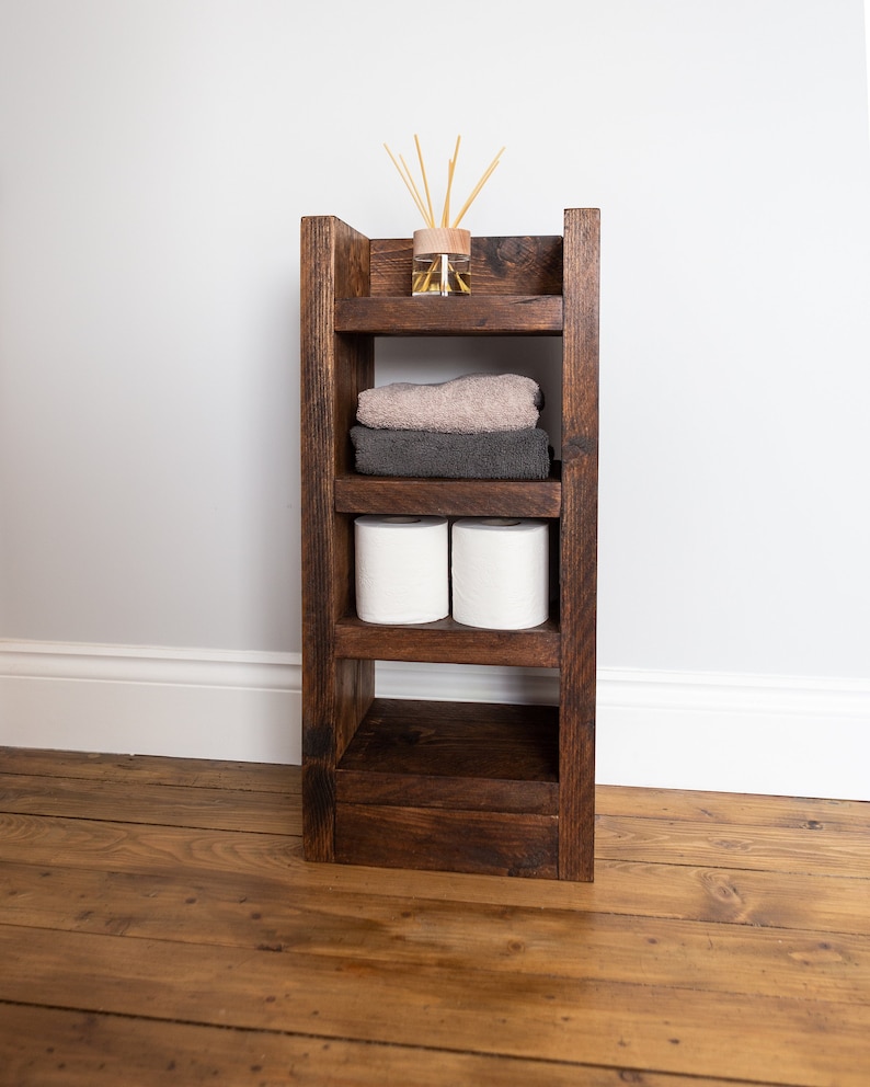 May include: A dark brown wooden shelf unit with four shelves. The top shelf has a glass jar with wooden sticks. The second shelf has two towels, one gray and one brown. The third shelf has two rolls of toilet paper. The bottom shelf is empty.