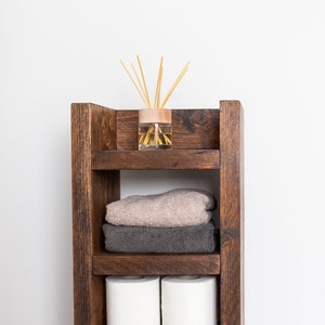 May include: A wooden shelf unit with three shelves, featuring a brown and beige towel, two rolls of toilet paper, and a glass diffuser with wooden sticks.