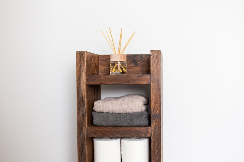 May include: A wooden shelf unit with three shelves, featuring a brown and beige towel, two rolls of toilet paper, and a glass diffuser with wooden sticks.