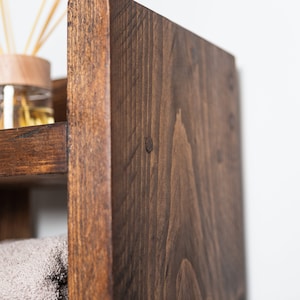 May include: A dark brown wooden shelf unit with three shelves. The top shelf has a glass jar with sticks in it. The bottom shelf has a towel on it.
