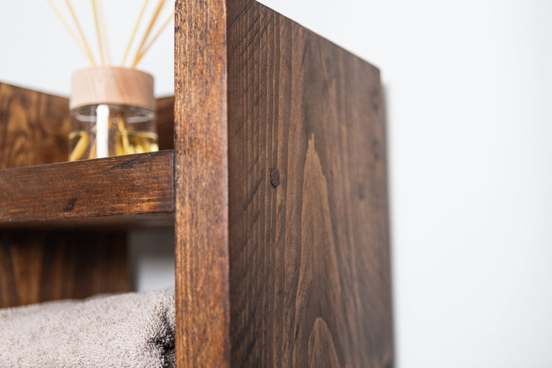 May include: A dark brown wooden shelf unit with three shelves. The top shelf has a glass jar with sticks in it. The bottom shelf has a towel on it.