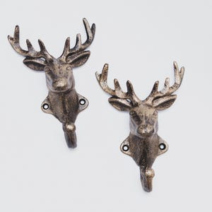 May include: Two gold-toned cast iron deer head hooks. The hooks are mounted on a white wall.