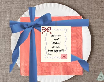 Puede incluir: Un plato de papel blanco con una servilleta coral está atado con una cinta azul. Una etiqueta con las palabras "dinner and dishes on us. bon appétit!" cuelga de la cinta. Un pequeño sobre rosa con un corazón también está en la etiqueta.