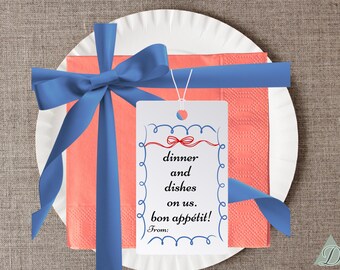 Puede incluir: Un plato de papel blanco con una servilleta coral y una cinta azul atada en un lazo. Una etiqueta blanca con el texto "dinner and dishes on us. bon appétit!" está adjunta a la servilleta.