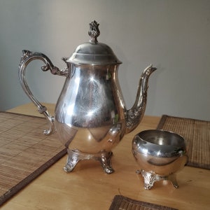 Puede incluir: Una tetera de plata con un diseño floral en la tapa y una jarra de crema a juego. La tetera tiene un cuerpo redondo y un pico curvo. La jarra de crema tiene un cuerpo redondo y una pequeña asa.