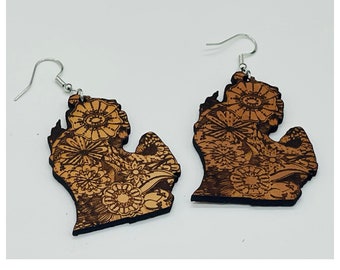 May include: A pair of wooden earrings shaped like the state of Michigan, with a floral design etched into the wood. The earrings are brown and have silver hooks.