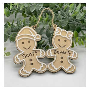 Può includere: Due decorazioni natalizie in legno a forma di omino di pan di zenzero. Uno indossa un cappello da Babbo Natale, l'altro un fiocco. I nomi "Scott" e "Beverly" sono stampati. Ogni ornamento ha un contorno bianco e uno spago di iuta per appenderlo.