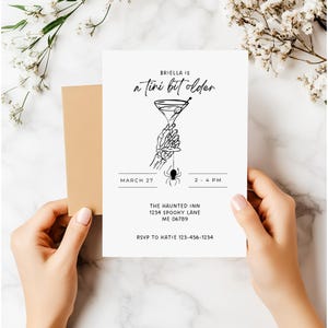 K&ouml;nnte beinhalten: Eine wei&szlig;e Einladungskarte mit schwarzem Text und einem beigefarbenen Umschlag. Auf der Karte steht "Briella is a tini bit older" mit einer Martini-Glas-Grafik, die von einer Skeletthand gehalten wird. Das Datum ist der 27. M&auml;rz, 14-16 Uhr.