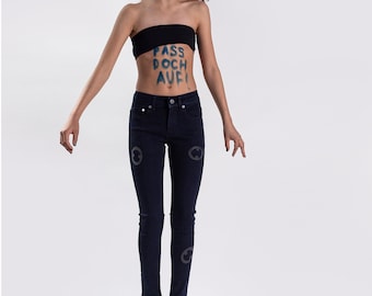 May include: A young person wearing a black strapless top and dark blue jeans with a message written on their stomach that says "Pass doch auf!"