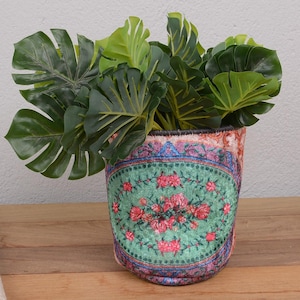 May include: A patterned plant pot featuring a floral design in pink, green, and blue. The pot holds a green plant with large leaves. The pot is placed on a wooden surface. The pot's design has a vintage feel.