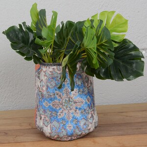 May include: A decorative vase with a blue and white floral pattern, holding vibrant green leafy plants. The vase has orange accents and sits on a wooden surface. The plants add a natural element to the decor.