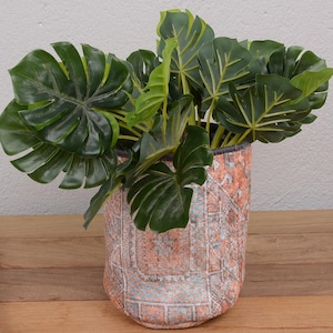 Könnte beinhalten: Eine dekorative Pflanze in einem strukturierten, zylindrischen Topf. Der Topf hat ein Muster aus Orange, Rosa und Grau. Die Pflanze hat große, grüne Blätter. Der Topf steht auf einer Holzoberfläche.
