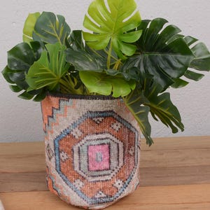 May include: A decorative plant pot made from a vintage rug, featuring a geometric pattern in shades of orange, pink, and blue. The pot is filled with lush, green Monstera leaves, creating a vibrant contrast. The pot sits on a wooden surface.