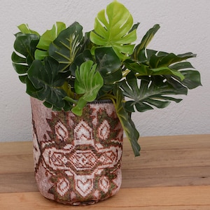 May include: A decorative plant pot with a brown and beige patterned design, filled with vibrant green leafy plants. The pot sits on a wooden surface, creating a natural and inviting aesthetic. The pot's design adds a touch of bohemian flair.