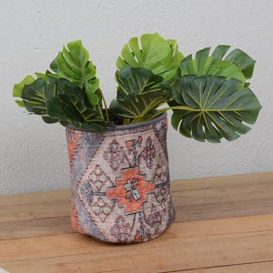 Peut inclure: Un pot en tissu décoratif avec un motif géométrique orange, gris et beige contient plusieurs grandes feuilles vertes. Le pot est posé sur une surface en bois, avec un mur neutre en arrière-plan.