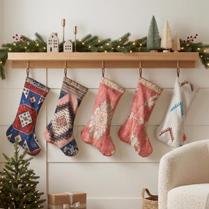 Puede incluir: Cinco calcetines navideños colgantes con diversos patrones tejidos en azul, rosa, rojo y blanco, suspendidos de una repisa de madera clara decorada con una guirnalda de pino, pequeñas casas de cerámica y árboles miniatura de cepillo.