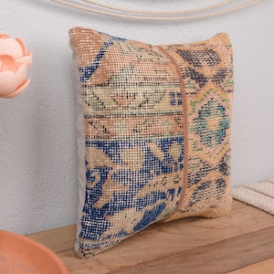 May include: A decorative pillow with a vintage rug design. The pillow features a patchwork of patterns in shades of blue, beige, and gold. A leather strip accents the center. The pillow is placed on a wooden surface.