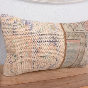 Puede incluir: Cojín rectangular decorativo con un diseño beige, azul y rosa. El cojín tiene un aspecto texturizado y un borde marrón. El cojín está colocado sobre una superficie de madera.