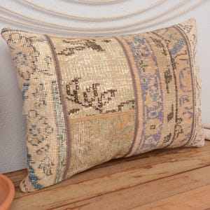 May include: Rectangular decorative pillow with a patchwork design. The pillow features a beige background with sections of blue, purple, and brown patterns. The pillow is placed on a wooden surface, with a vase of flowers and other decor in the background.