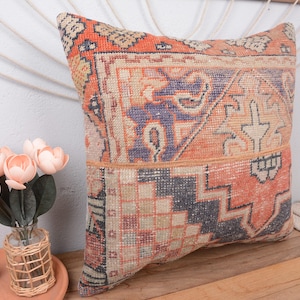 May include: Square pillow with a vintage rug design in shades of orange, beige, and navy blue. The pillow has a patchwork style with geometric patterns. A vase of pink flowers sits nearby.