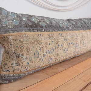 May include: Rectangular decorative pillow with a vintage rug design. The pillow features a gray top section with blue floral patterns and a beige bottom section with intricate floral and geometric motifs. The pillow is placed on a wooden surface.