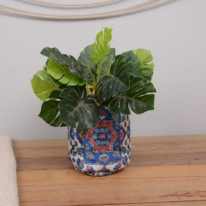 May include: A decorative ceramic pot with a blue, red, and white pattern holds a vibrant green plant with large, split leaves. The pot sits on a wooden surface, with a neutral-colored fabric partially visible on the left.