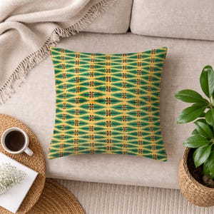 Puede incluir: Cojín decorativo cuadrado con un patrón repetitivo de formas verdes y marrones sobre un fondo amarillo. El cojín está colocado sobre un sofá beige, con una taza de café y una planta visibles en el fondo.