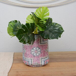 Op de afbeelding: Een decoratieve plantenbak met een roze, grijze en witte patroon bevat levendige groene kunstmatige Monstera-bladeren. De plantenbak staat op een houten oppervlak. De plantenbak is ongeveer 15 cm hoog.